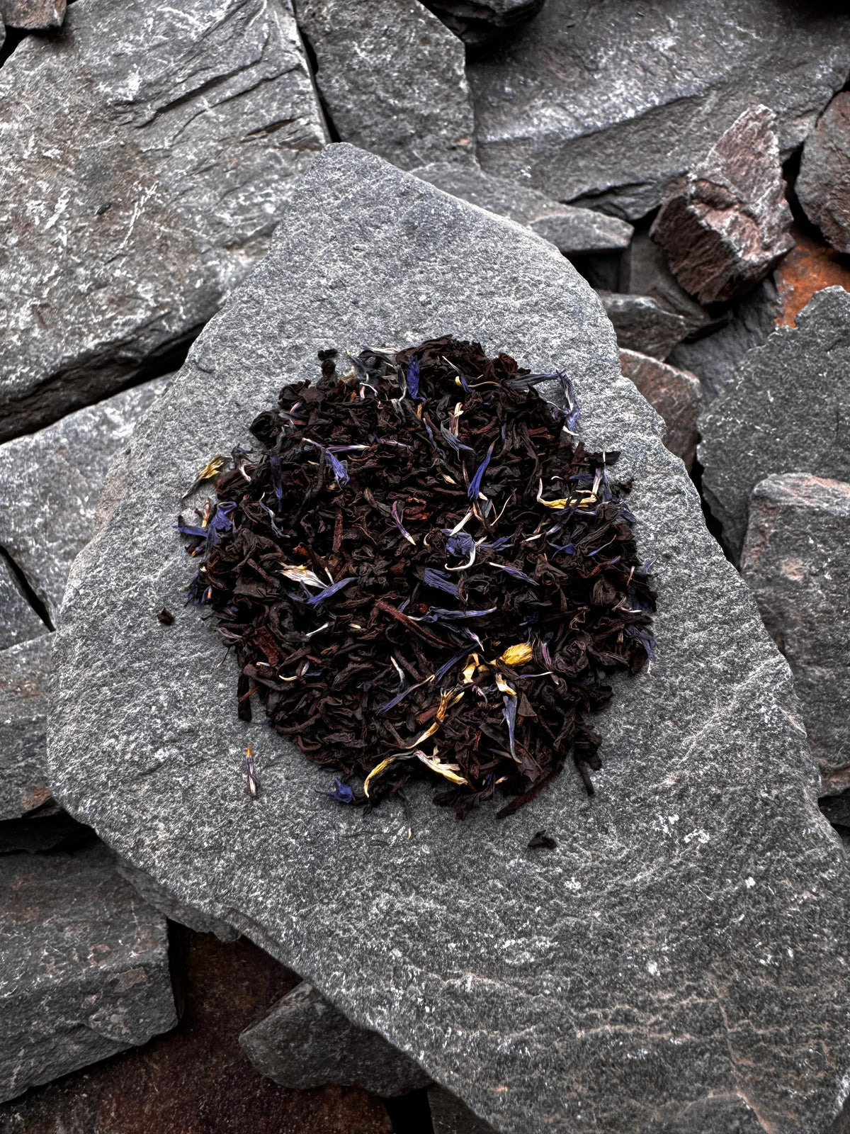 A small mound of High Summit Tea Co.’s Earl Grey loose leaf tea, with blue petals visible, on a grey slate slab surrounded by slate pieces